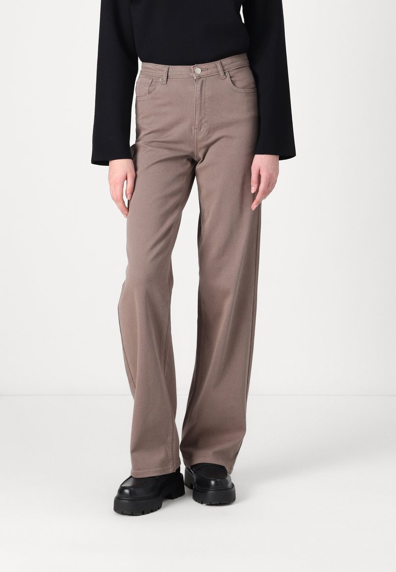 Personne portant un pantalon taupe taille haute à jambes larges, un haut noir à manches longues et des chaussures noires à semelle épaisse, debout sur un fond blanc uni.