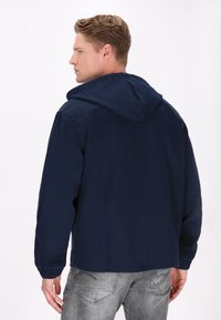 Marineblå hættetrøje med elastiske ærmer, der har en glat tekstur og et understated design. Båret med grå jeans, set fra ryggen.