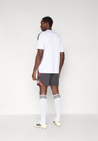 Sportbekleidung mit weißem kurzärmligem Shirt, grauer Shorts mit roten Akzenten, weißen knielangen Socken mit schwarzen Streifen und Stollenschuhen.