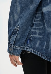 Roka ar sudraba gredzenu blakus zilas džinsa jaku aprocei ar "HUGO" uzrakstu, vilkta pār melnām biksēm ar mežģīņu detaļu.