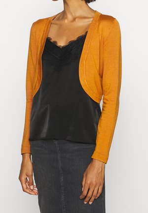 Femme portant un caraco noir bordé de dentelle sous un cardigan court couleur orange brûlé à manches longues et une jupe en denim foncé.