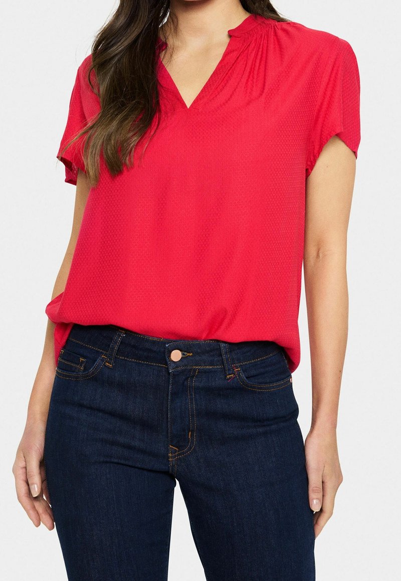 Femme portant un chemisier rouge vif à manches courtes rentré dans un jean bleu foncé, avec de longs cheveux bruns sur une épaule.