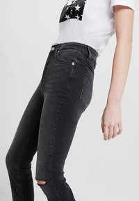 Vaqueros negros de tiro alto, ajustados, con un detalle de rodilla rasgada, un desvanecimiento sutil y un estilo clásico de cinco bolsillos.