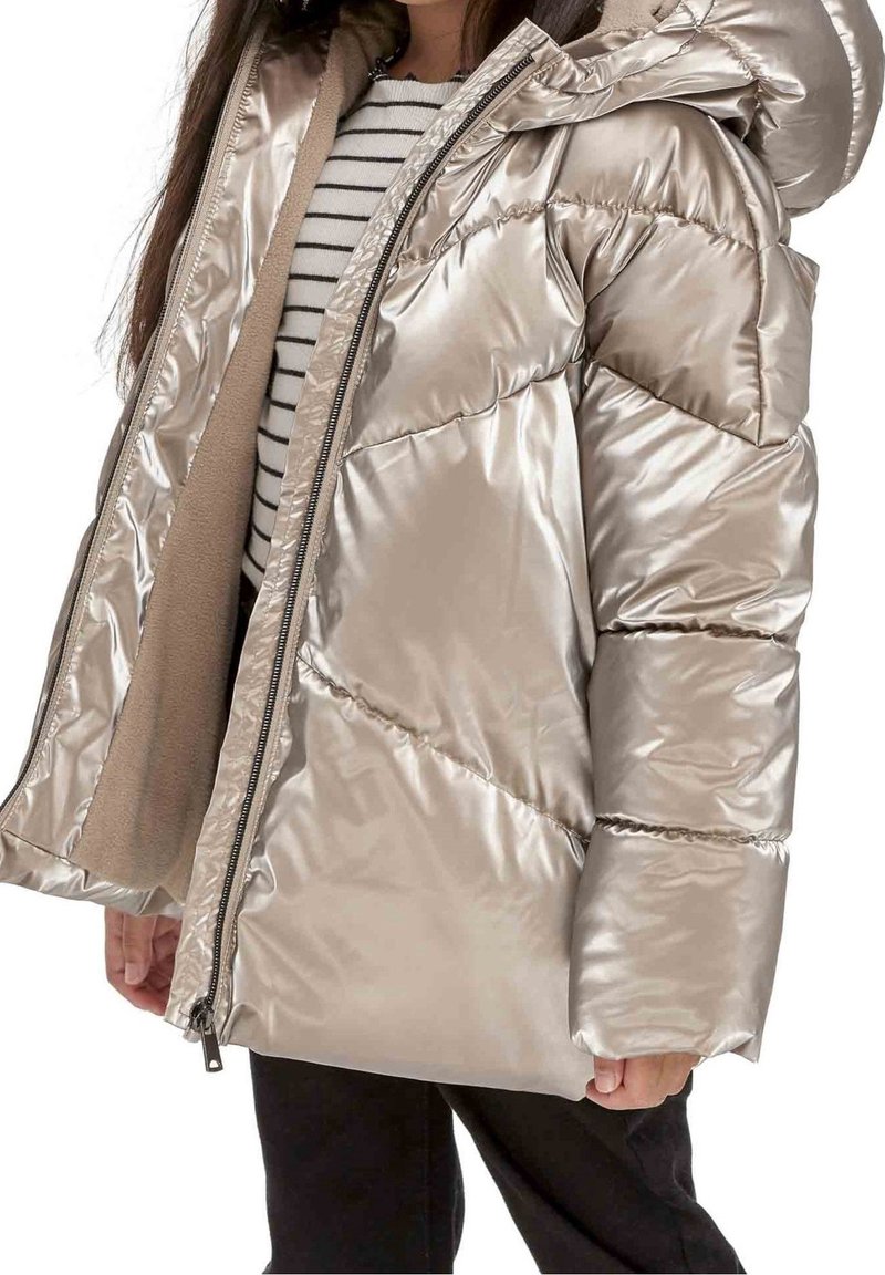Enfant portant une doudoune beige métallique partiellement dézippée avec capuche, chemise rayée en dessous et pantalon noir.