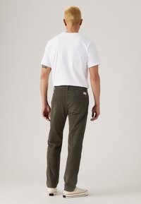 Man with short blonde hair wearing a white t-shirt, dark green pants, and white sneakers, standing facing away against a plain background.