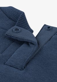 Tmavomodrá texturovaná pletená látka s polo golierom. Má dva gombíky a rebrované detaily na golieri a ramenách.