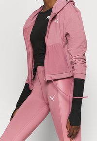 Frau in rosa Puma-Reißverschlussjacke und passenden Leggings, mit schwarzem Puma-Langarmshirt darunter, steht vor einem weißen Hintergrund.