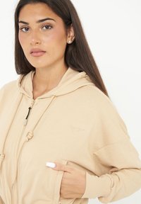 Sudadera beige con cremallera, capucha ajustable, bolsillos frontales y puños acanalados. Presenta un pequeño logo en la parte superior izquierda. Textura de tela suave.