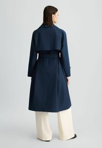 Cappotto trench navy con alette posteriori, vita con cintura e maniche lunghe. Tessuto liscio, design fino al ginocchio, abbinato a pantaloni larghi di colore crema.