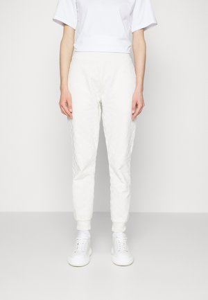 Person iført teksturerede hvide joggers, hvide sneakers og en enkel hvid kortærmet skjorte står foran en hvid baggrund.