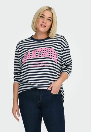 Lyshåret kvinde iført navy- og hvidstribet langærmet skjorte med lyserød tekst "Santorini Yacht Club Summer of Love" og mørke jeans.