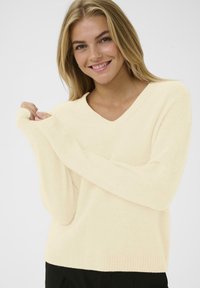 Kremfärgad V-ringad tröja i mjukt stickat material, med långa ärmar och ribbad nederkant, bärs på ett avslappnat sätt med loose fit.