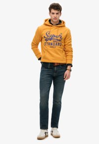 Felpa giallo con testo blu, jeans in denim scuro e sneakers bianche con accenti grigi. La felpa presenta una tasca a marsupio e cappuccio con cordini.