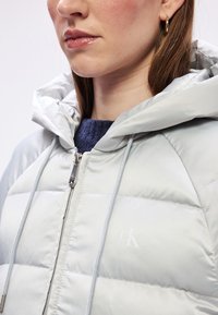 Svijetlosivi puffa jakna s kapuljačom, koja ima zatvaranje na zip, vrpce za podešavanje i kviltani uzorak. Sadrži suptilni logo na prsima.