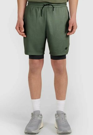 Mężczyzna noszący oliwkowo-zielone sportowe spodenki z czarnymi legginsami kompresyjnymi pod spodem, białe skarpetki za kostkę i szare buty do biegania.