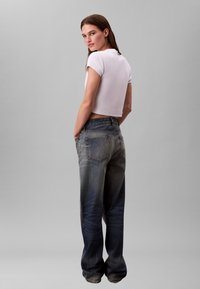 Lühendatud valge t-särk, mida on kombineeritud laia säärega tuhmunud siniste teksapükstega, millel on viis taskut ja veidi kulunud viimistlus.