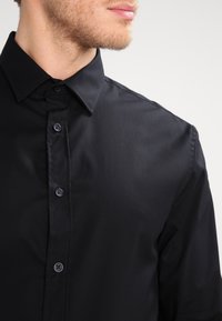 Zwarte button-up shirt gemaakt van gladde stof, met een puntige kraag en donkere knopen langs de sluiting. Eenvoudig en klassiek ontwerp.