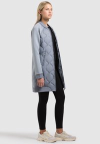 Nő világoskék hosszú, steppelt kabátban, fekete leggingsben és világos bézs sportcipőben áll egyszínű szürke háttér előtt.