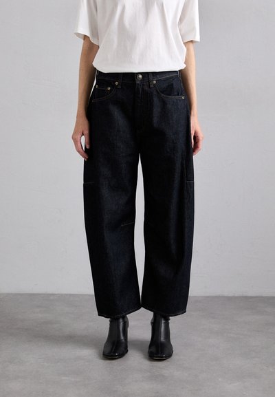 Donkerdenim wijde broek met voorzakken, sluiting met bovenste knoop en zichtbare stiksels. G gedragen met een witte t-shirt en zwarte laarzen.