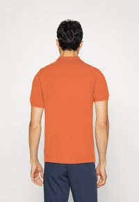 Oranžová polo košile s krátkým rukávem a úzkým límečkem. Má měkkou strukturu a přiléhavý design, doplněná tmavě modrými kalhotami.