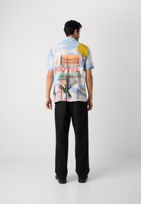 Majica s kratkimi rokavi v večbarvnem dizajnu, ki prikazuje slikovit potniški motel z palmami, soncem in živahnimi napisi na modri podlagi.