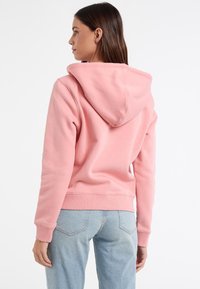 Felpa rosa con cappuccio a coulisse, polsini e orlo a costine, e una texture morbida. Indossata con jeans azzurri chiari, leggermente strappati.