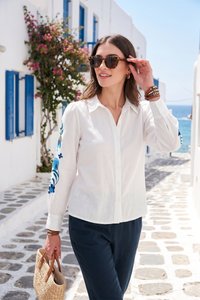 Woman wearing sunglasses, white embroidered blouse, and dark pants holds a straw bag, walking on a sunlit stone street by the sea.
