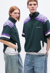 Πράσινο σακάκι με φερμουάρ, με μοτίβα σε γεωμετρικό σχήμα μωβ στο ύψος των ώμων, λευκές λεπτομέρειες στο τελείωμα και άνετη εφαρμογή. Φοριέται με ανοιχτό μπλε τζιν.