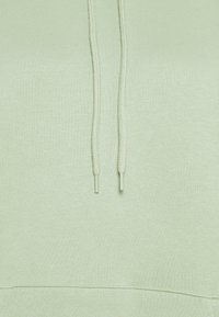Světlé zelená mikina s kapucí z měkké látky, vybavená stahovací kapucí a klokaní kapsou v oblasti pasu. Jemný texturovaný detail je viditelný.