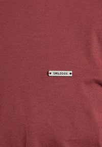 T-shirt bordeaux realizzata in tessuto morbido; presenta un'etichetta grigia rettangolare con la scritta "SMILODOX" attaccata vicino all'orlo.