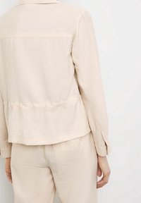 Jasno beżowa, teksturowana kurtka o luźnym kroju, z długimi rękawami i przednim karczkiem. Posiada taliowanie i boczne rozcięcia.