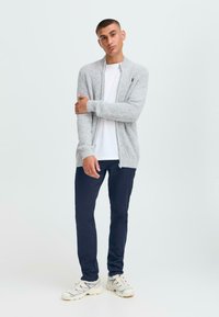 Grå strikket cardigan med glidelås, lange ermer, ribbestrikket mansjetter og et subtilt mønster, kombinert med en hvit t-skjorte og mørke bukser.