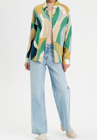Personne portant une chemise à motifs abstraits colorés, un jean large bleu clair, un pull à col roulé beige et des chaussures beiges sur fond blanc.