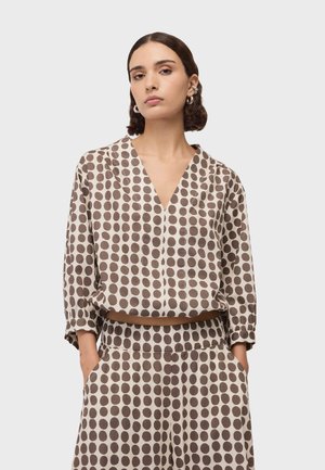 Femme portant un haut et un pantalon beige à gros pois marron, debout les mains dans les poches devant un fond uni.