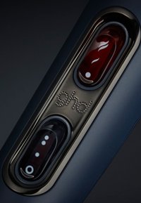 Outil capillaire Ghd avec un corps en bleu mat foncé, comportant deux boutons de couleur : un rouge et un noir, avec un logo argenté et un motif de points.
