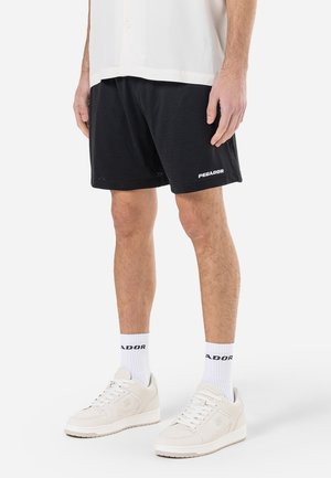 Pantalones cortos deportivos de malla negra con el logo blanco "PECADOR", combinados con zapatillas y calcetines blancos que cuentan con el texto negro "PECADOR".