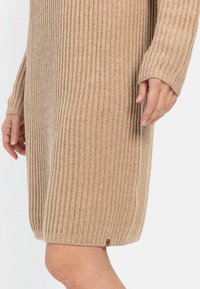 Beiges Strickkleid mit vertikaler Rippung, langen Ärmeln und einem leicht gewölbten Saum; verfügt über ein kleines Markenetikett in der Nähe der unteren Naht.