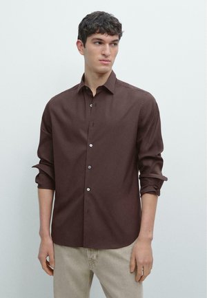 Giovane uomo con capelli corti e scuri che indossa una camicia marrone scuro a maniche arrotolate e pantaloni beige chiaro, che guarda verso sinistra.