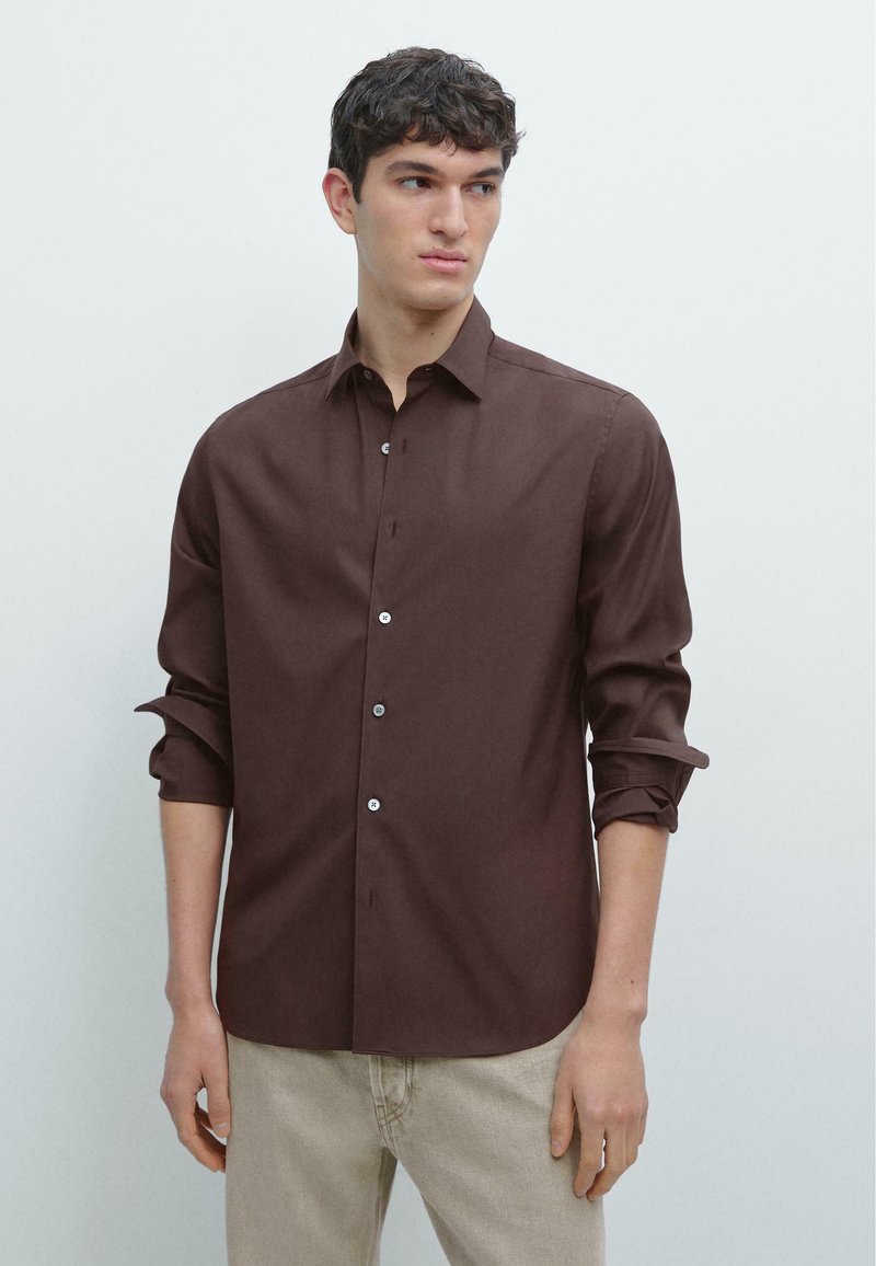 Jeune homme aux cheveux courts et foncés, portant une chemise marron foncé à manches retroussées et boutonnée, ainsi qu'un pantalon beige clair, regardant vers sa gauche.