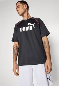 Maillot de sport noir avec des accents rouges, à manches courtes, arborant un grand logo PUMA blanc. Porté avec un short blanc, avec des détails d'étiquette sur le short.