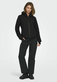 Veste noire imperméable avec capuche, fermeture éclair à l'avant et poches latérales, associée à un pantalon noir et des chaussures noires robustes.