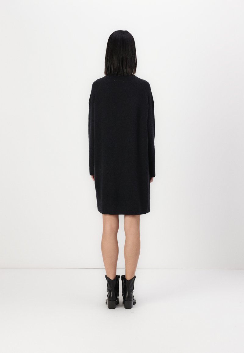 Schwarzes Strickkleid mit langen Ärmeln, lässiger Passform und mittellanger Länge bis zum Oberschenkel. Kombiniert mit schwarzen Ankle Boots, vor einem weißen Hintergrund präsentiert.