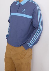 Modrá polo košile s dlouhým rukávem a zipovým límcem, s světle modrým vodorovným pruhem a třemi modrými pruhy na každém rukávu. Logo Adidas na hrudi.