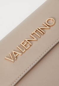 Bežs ādas maks ar izsistām "VALENTINO" zīmēm zeltainā krāsā, ar redzamām šuvēm pa malām, kas piešķir papildu detalizāciju.