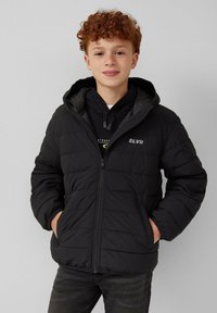 Zwarte pufferjack met een rits aan de voorkant, gewatteerde textuur en een capuchon. Kenmerkt zich door een logo op de borst en zijzakken. Draag het over een zwarte hoodie.