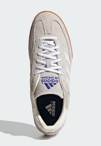 Šedé semišové tenisky s bílými tkaničkami, se modrým brandingem Adidas a černou stélkou. Podešev z gumy. Klasický design a nízký profil.