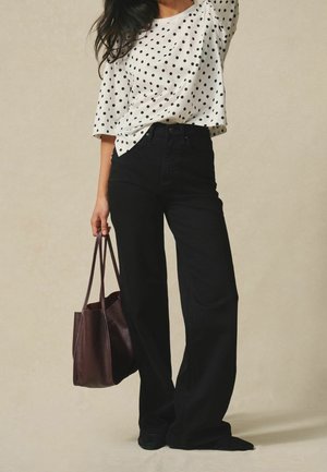 Femme portant un top blanc à pois et un pantalon large noir, tenant un sac cabas en cuir brun foncé à ses côtés.
