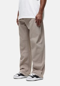 Man in beige wijde broek, witte T-shirt en zwart-witte sneakers, staand tegen een effen lichte achtergrond.
