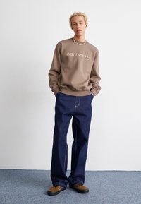 Barna pulóver fehér "Carhartt" logóval, tengerészkék széles szárú farmerkabát és barna cipő párosításában. A modell egy sima háttér előtt áll.