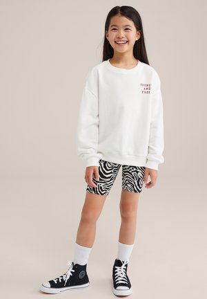 Vit sweatshirt med texten "YOUNG AND FREE" i rött, i kombination med cykelshorts med svart och vit zebramönster samt svarta högskurna sneakers.